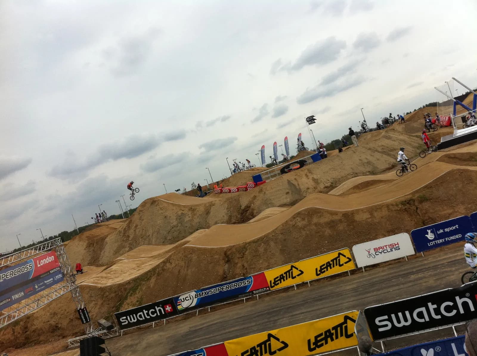 A BMX rider in a red shirt and helmet catches air over a dirt jump on a cloudy day, with other riders and spectators visible on the track and surrounding hills.